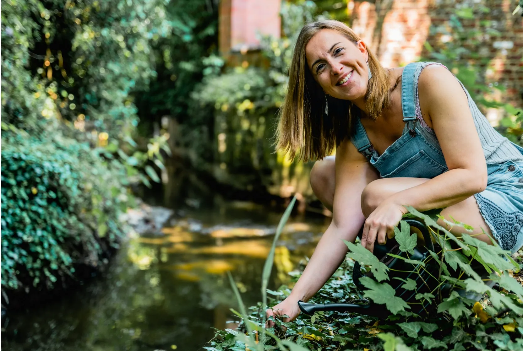 Een vrouw in een denim tuinbroek zittend naast een kabbelende beek in een weelderige tuin. Ze glimlacht en lijkt te genieten van de natuur terwijl ze zich over de planten buigt.