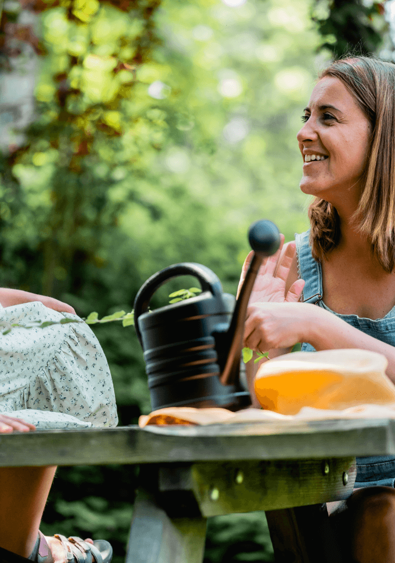 Close-up van een vrolijke vrouw met een gieter, zittend naast een houten tafel met een kruik en wat eten, omgeven door een weelderige groene omgeving.
