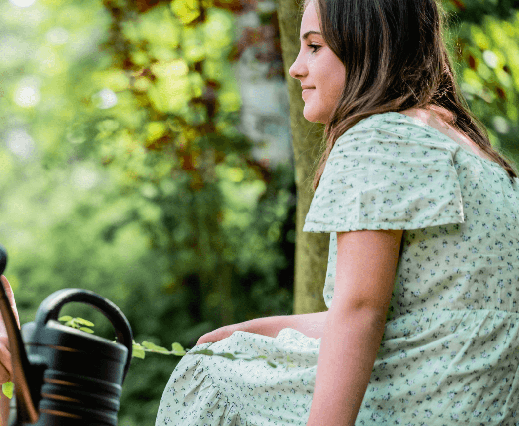 Zijprofiel van een jonge vrouw in een jurk met patroon, peinzend zittend te midden van een serene groene achtergrond.