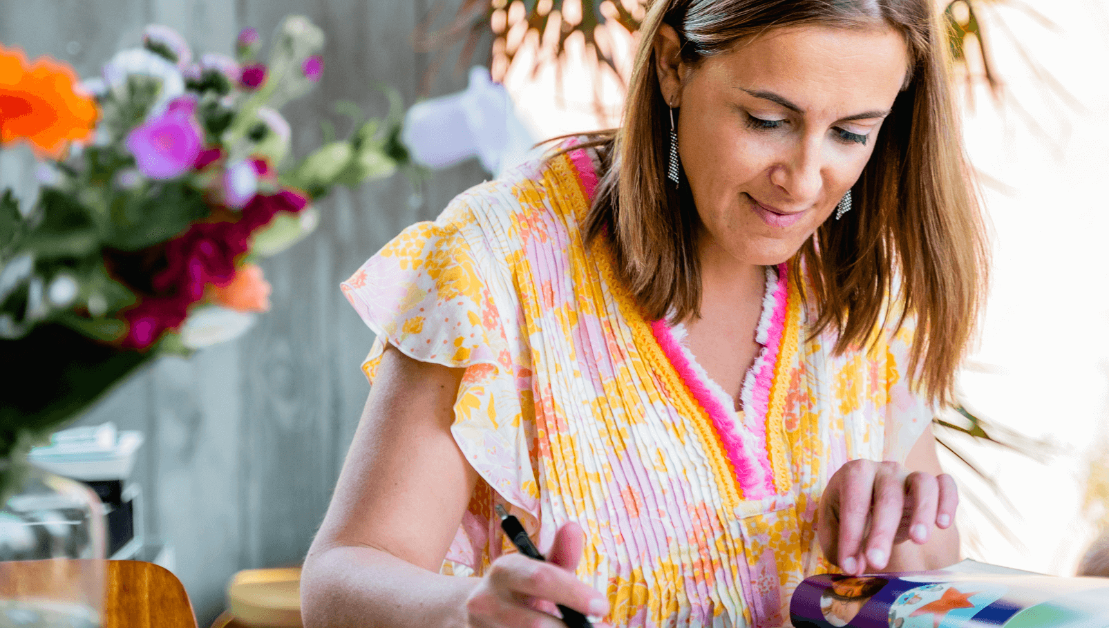 Een gefocuste vrouw in een jurk met levendige patronen, die schrijft of tekent in een kleurrijk dagboek, gelegen naast een boeket verse bloemen met een rustieke houten achtergrond.
