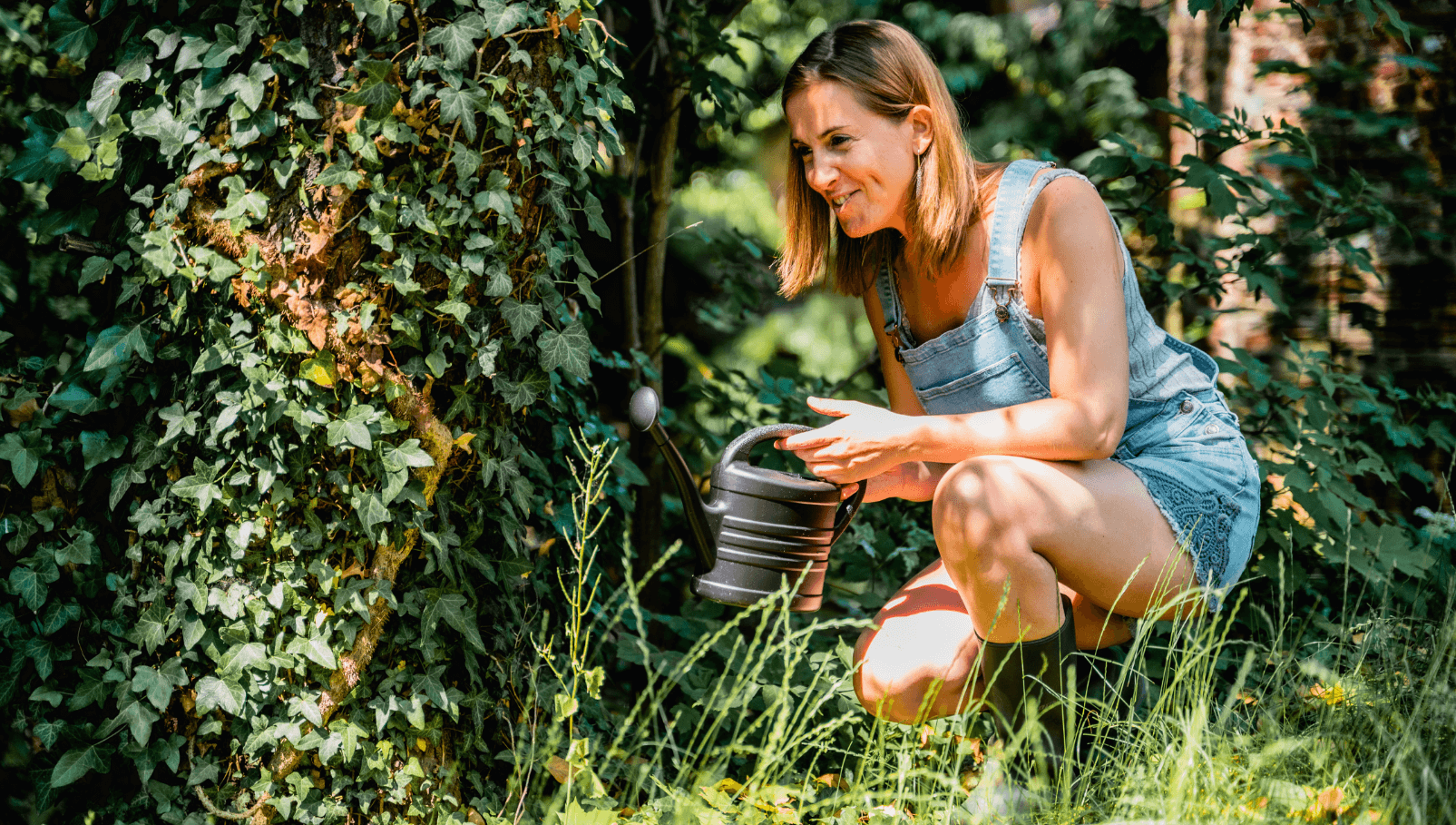 Een vrouw in spijkeroverall knielt in een weelderige tuin en geeft de planten voorzichtig water met een kleine gieter, omringd door levendig groen en met klimop bedekte bomen.