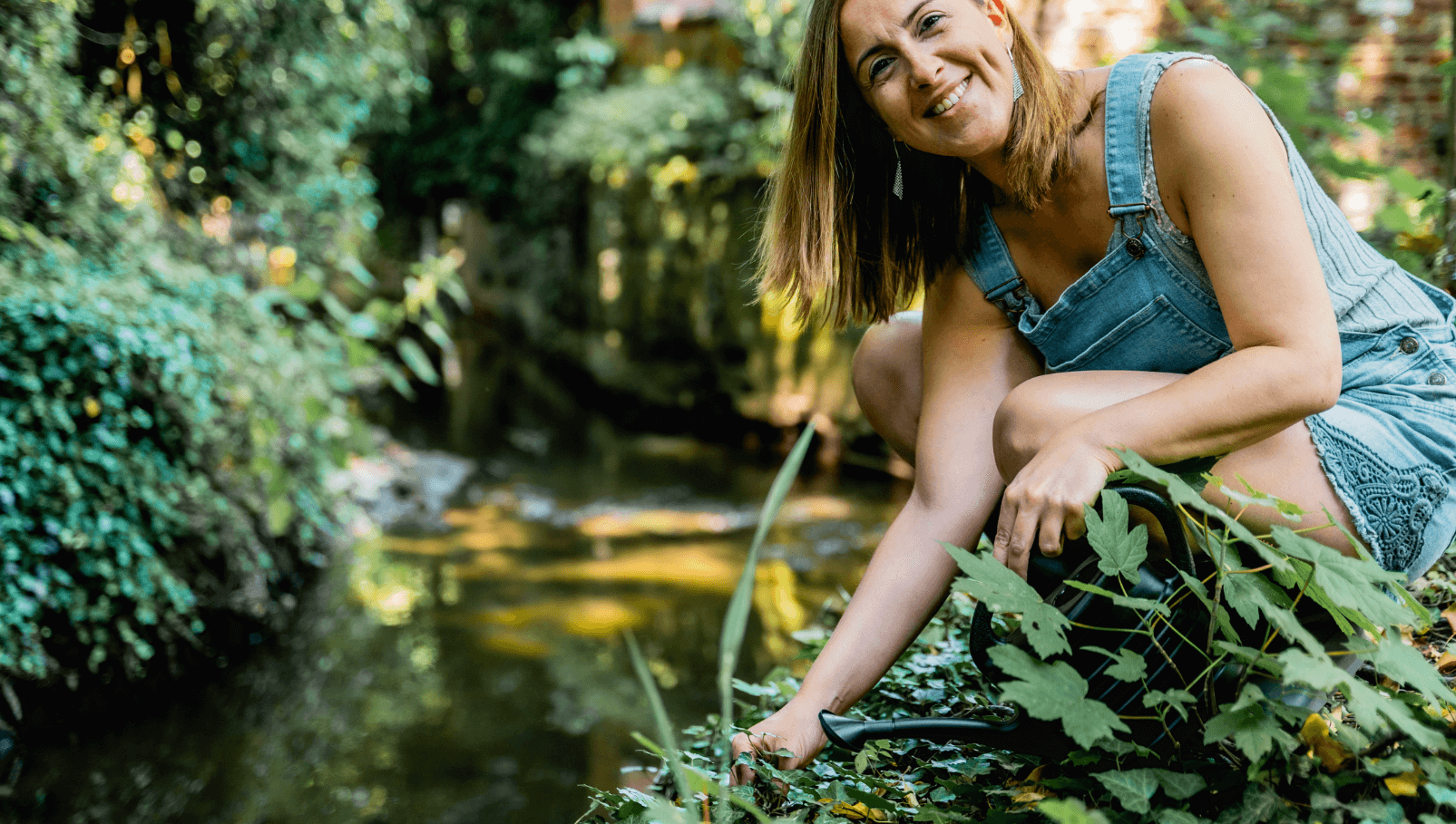 Een vrouw in een denim tuinbroek zittend naast een kabbelende beek in een weelderige tuin. Ze glimlacht en lijkt te genieten van de natuur terwijl ze zich over de planten buigt.